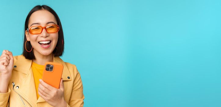 Happy stylish asian girl using smartphone and laughing, smiling at camera, standing over blue background. Copy space