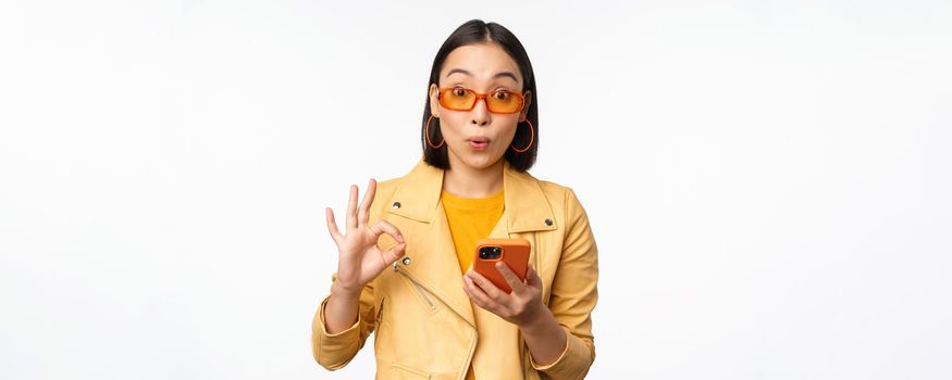 Amazed asian girl in sunglasses, showing okay, ok sign, holding smartphone, looking impressed, recommending smth, standing over white background.