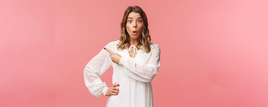 Fascinated and excited, curious blond caucasian girl in white dress, look intrigued at camera with folded lips, say wow and pointing finger left at blank space, standing pink background.