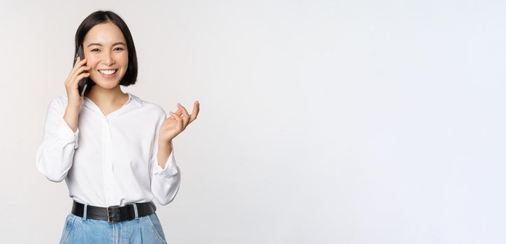 Smiling happy asian woman talking on smartphone with client, saleswoman on call, holding mobile phone and gesturing, standing over white background.
