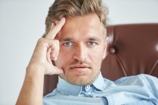 Portrait of a stylish intelligent man, small unshaven, charismatic, blue shirt, sitting on a brown leather chair, dialog, negotiation, short sleeve, brutal, hairstyle, emotions, energy, interest. High quality photo