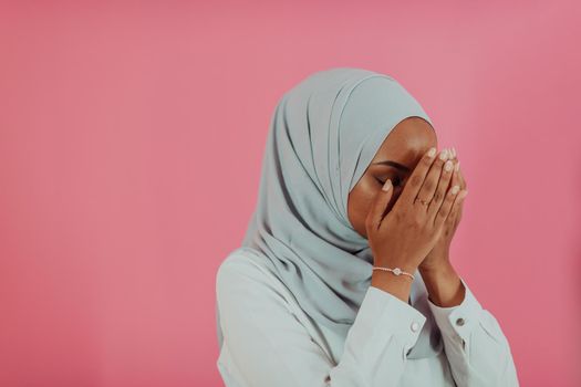 Modern African Muslim woman makes traditional prayer to God, keeps hands in praying gesture, wears traditional white clothes, has serious facial expression, isolated over plastic pink background. High-quality photo