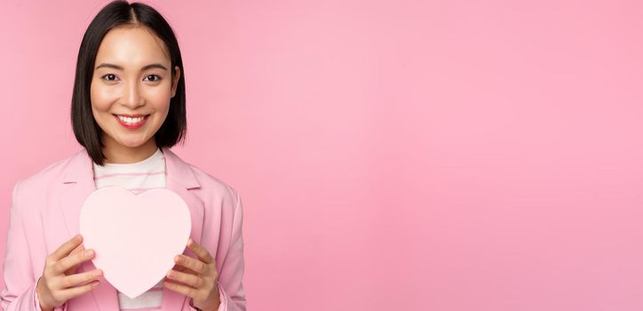 This is for you. Romantic cute asian corporate woman, girl in suit, showing heart-shaped box with gift, standing over pink background.