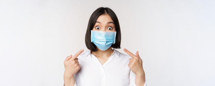 Image of amazed young asian woman pointing at herself while wearing medical face mask, standing over white background.