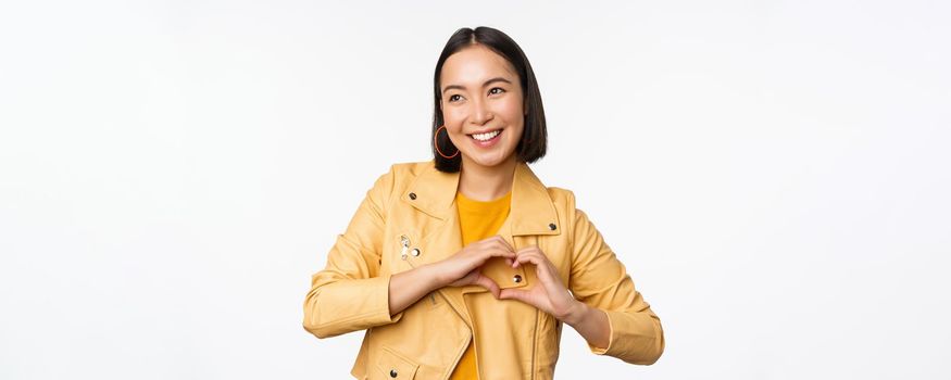 Image of beautiful asian girl showing heart, love sign, express care and romantic feelings, standing over white studio background.