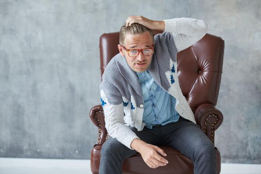 Portrait of a stylish intelligent man with glasses stares into the camera, good view, small unshaven, charismatic, blue shirt, gray sweater, sitting on a brown leather chair, grabs his head. High quality photo