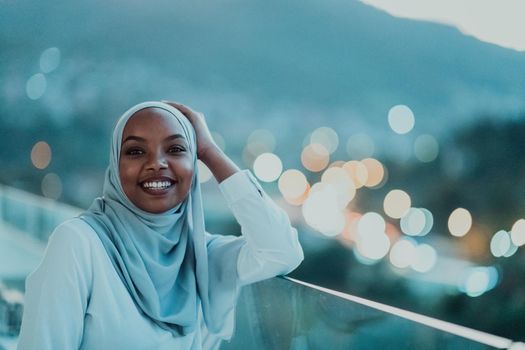 African Muslim woman in the night on a balcony smiling at the camera with city bokeh lights in the background. High-quality photo