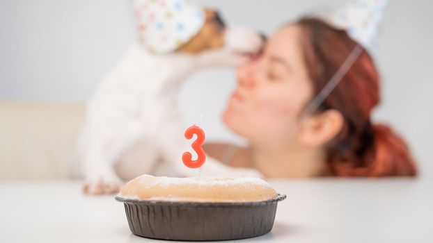 Caucasian woman and jack russell terrier in holiday caps look at the cake with a candle. The dog and the owner celebrate the third birthday