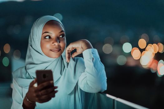 Young Muslim woman wearing scarf veil on urban city street at night texting on a smartphone with bokeh city light in the background. High-quality photo