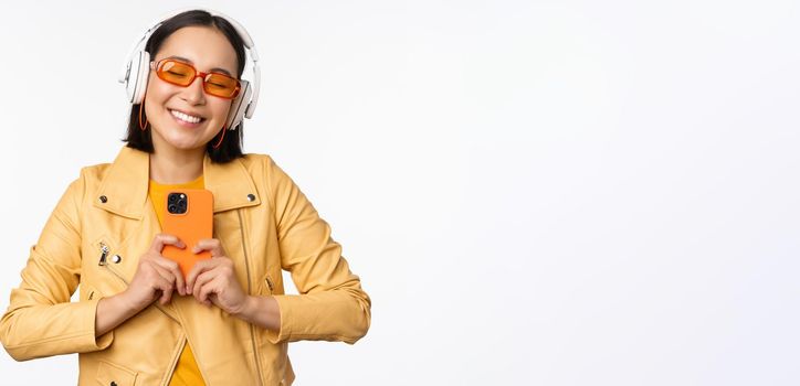 Technology concept. Stylish asian girl in headphones, holding smartphone, dancing and singing, listening music, standing over white background.