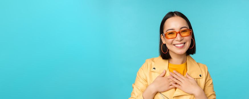Close up portrait of asian young woman in sunglasses, smiling and looking romantic, standing happy over blue background. Copy space