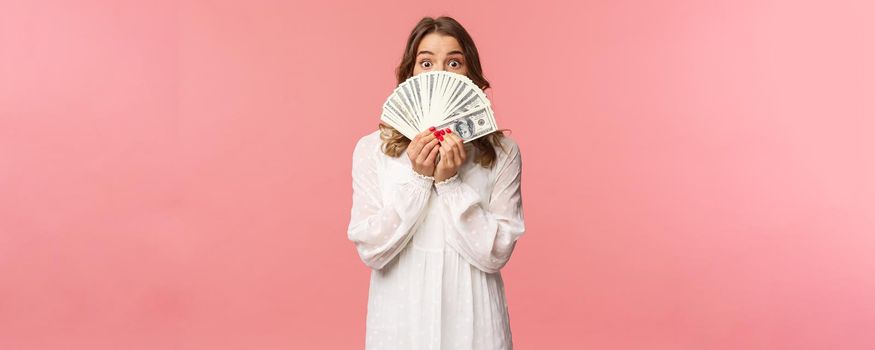 Portrait of amazed and excited cute feminine blond girl in white dress, holding dollars over face, looking from under cash at camera with surprised expression, stand pink background.