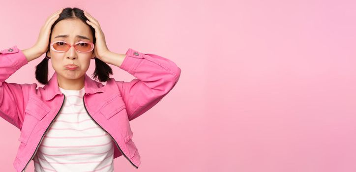 Portrait of troubled asian woman, korean girl holds hands on head and look in panic, standing distressed and shocked against pink background.