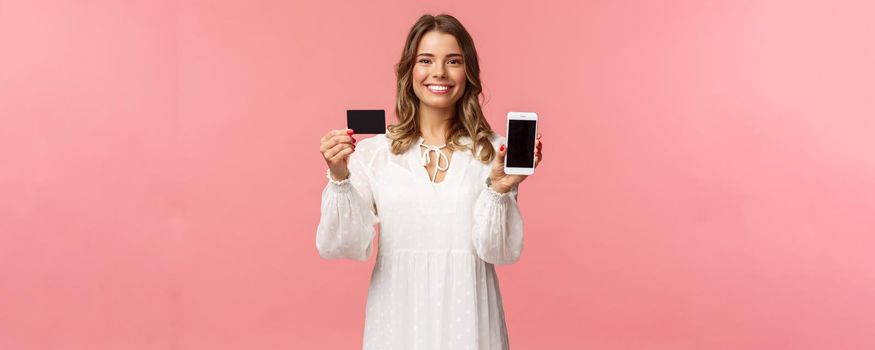 Portrait of satisfied cute blond girl in white dress, showing you her credit card and mobile phone application, online shopping app or bank website, smiling camera, standing pink background.