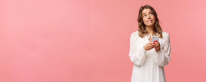 Portrait of dreamy and thoughtful girl thinking what order, using application to make reservation, look up and bite lip pondering, thinking as using mobile phone over pink background.