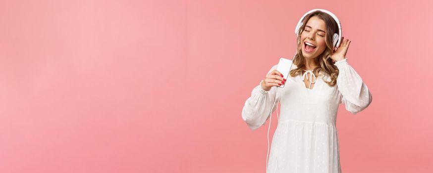 Portrait of beautiful blond caucasian female in white dress, listening music in headphones, close eyes and smiling, using mobile phone like microphone, singing along favorite song, pink background.