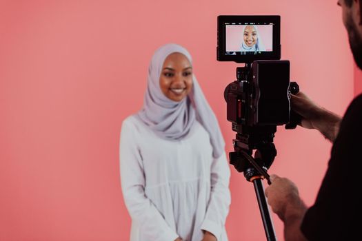 Videographer in digital studio recording video on professional camera by shooting female Muslim woman wearing hijab scarf plastic pink background. High-quality photo