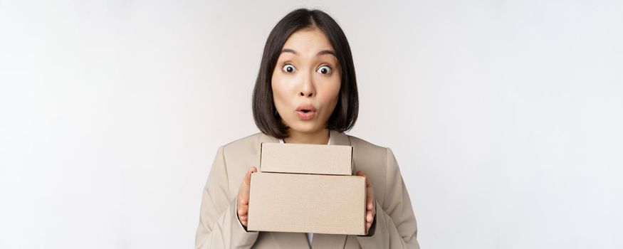 Portrait of asian saleswoman looking surprised, holding boxes, delivery goods, standing amazed in suit against white background.