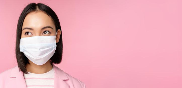 Close up portrait of japanese corporate woman in medical face mask from covid-19, looking left at logo, sale promo, standing over pink background.