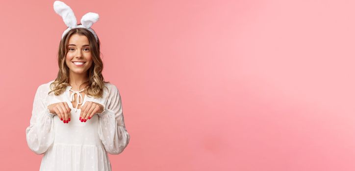 Holidays, spring and party concept. Portrait of cute and tender, lovely blond girl imitating bunny, holding hands like paws and wearing rabbit ears, smiling camera, pink background.