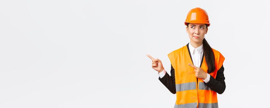 Building, construction and industrial concept. Indecisive asian female architect being unsure, wearing reflective clothing and helmer, smirk as pointing and looking upper left corner puzzled.