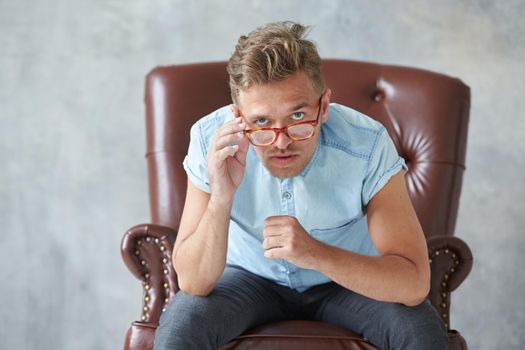 Portrait of a stylish man with glasses, stares into the camera, unshaven, charismatic, blue shirt with short sleeve, sitting on a brown leather chair, dialogue, negotiation, lower a little glasses. High quality photo