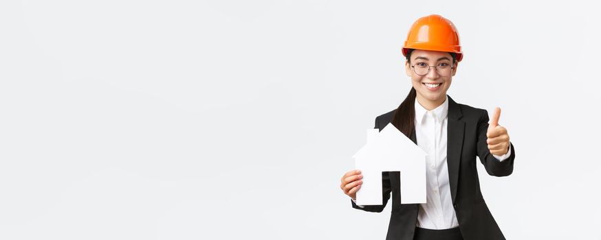 Portrait of young asian female construction engineer, architect in safety helmet and business suit holding home maket and show thumbs-up, promoting company service of building houses.