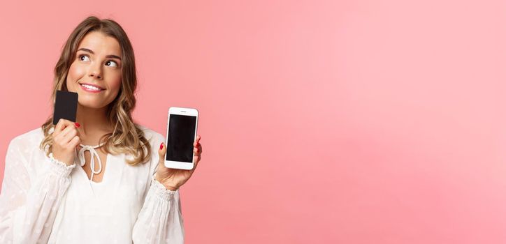 Finance, shopping and technology concept. Close-up portrait of thoughtful, dreamy attractive blond girl in white dress, thinking what buy, order something online shop, hold smartphone and credit card.