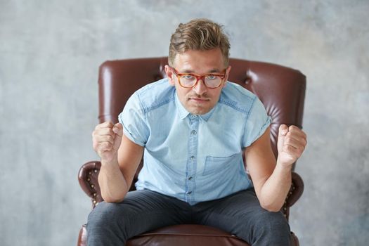 Portrait of a stylish intelligent man stares into the camera, small unshaven, charismatic, blue shirt, sitting on a brown leather chair, dialog, negotiation, short sleeve, brutal, hairstyle. High quality photo