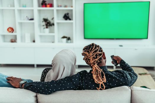 African Couple Sitting On Sofa Watching TV Together Chroma Green Screen Woman Wearing Islamic Hijab Clothes. High quality photo