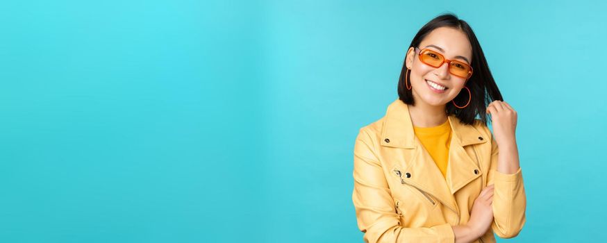 Young stylish asian woman in sunglasses smiling, playing with her haircut and looking happy, posing against blue background.