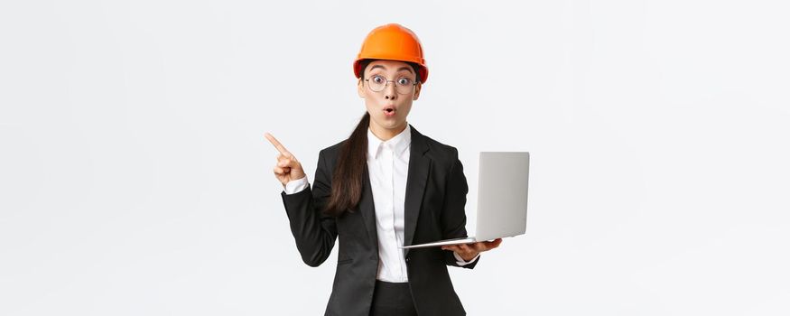 Impressed asian female entrepreneur at factory wearing safety helmet and business suit, pointing finger left and holding laptop computer, showing diagram at enterprise, white background.