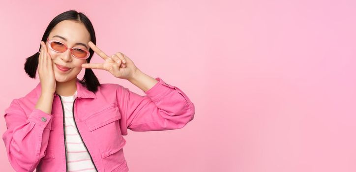 Portrait of kawaii asian girl in sunglasses, showing peace, v-sign near eye and smiles cute at camera, posing in trendy clothes against pink background.