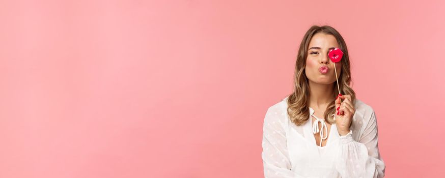 Spring, happiness and celebration concept. Close-up portrait of feminine lovely blond girl in white dress, folding lips in kiss and holding carton stick, partying having fun, stand pink background.