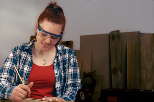 Young woman carpenter smilling, with red hair, working on wood design in a small carpentry workshop, dressed in blue checked shirt and red t-shirt. Young businesswoman handcrafting a piece of wood in her small business. Warm light indoors, background with wooden slats. Horizntal.