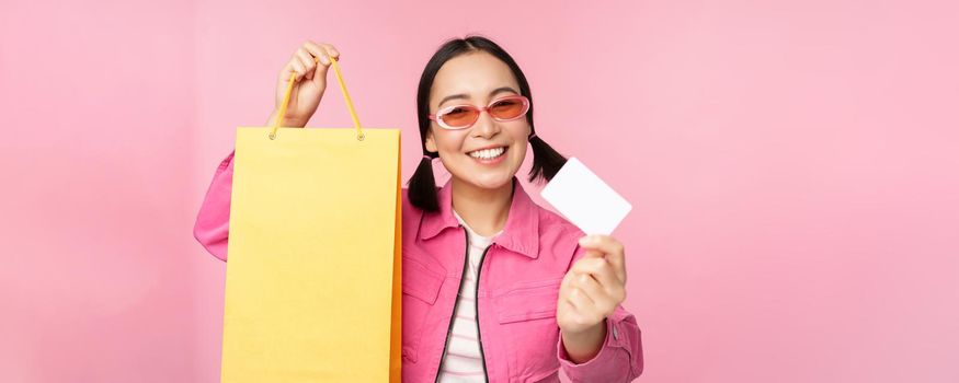 Happy young asian woman showing credit card for shopping, holding bag, buying on sale, going to the shop, store, standing over pink background.