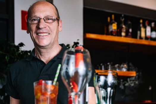 Male waiter, mature, bald, smiling, dressed in company uniform, black polo shirt, mature, experienced, holding hands on tray. Worker in his small business carrying a tray of drinks. Warm atmosphere and dim lighting. Horizontal