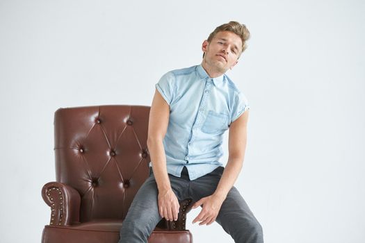 Portrait of a stylish intelligent man, small unshaven, charismatic, blue shirt, sitting on a brown leather chair, dialog, negotiation, short sleeve, brutal, hairstyle, emotions, indifference, fatigue. High quality photo