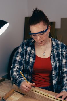 Young carpenter with red hair, measuring a strip of wood in her small carpentry shop, dressed in blue checked shirt and red t-shirt. Young businesswoman handcrafting a piece of wood in her small business. Warm light indoors, background with wooden slats. Vertical