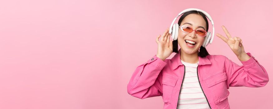 Dancing stylish asian girl listening music in headphones, posing against pink background.