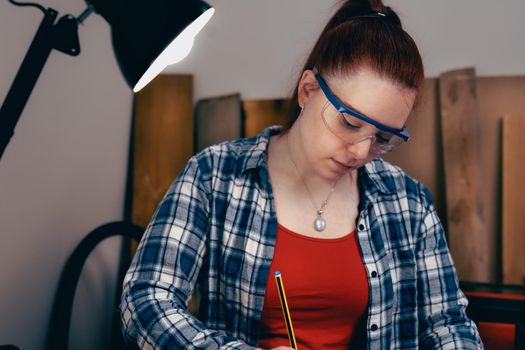 Young carpenter with red hair, working on wood design in a small carpentry workshop, dressed in blue checked shirt and red t-shirt. Young businesswoman handcrafting a piece of wood in her small business. Warm light indoors, background with wooden slats. Horizntal.