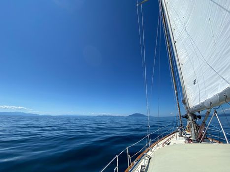 View of sailboat sailing along with sails up and tilted by the wind. Blue skies and mountains in the foreground