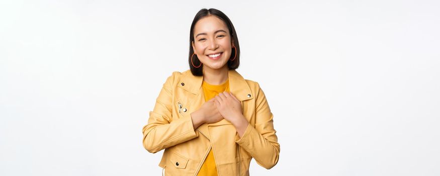 Portrait of smiling, beautiful asian girl looking with appreciation, thank you gesture, holding hands on heart flattered, standing over white background.