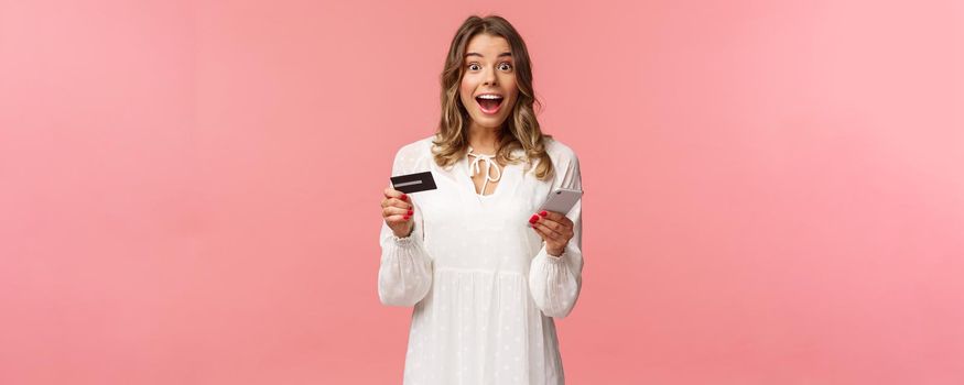 Portrait of excited and amused, happy smiling caucasian woman making order internet, shopping online with mobile phone, enter credit card, billing info, look camera amazed, pink background.