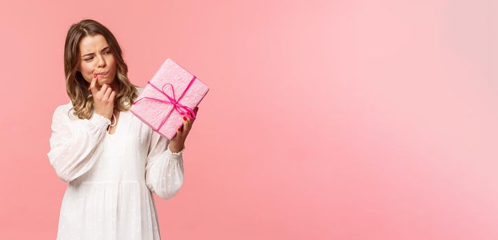Holidays, celebration and women concept. Portrait of thoughtful curious blond girl thinking whats inside gift box, look pondering say hmm as making assumpion, standing pink background.