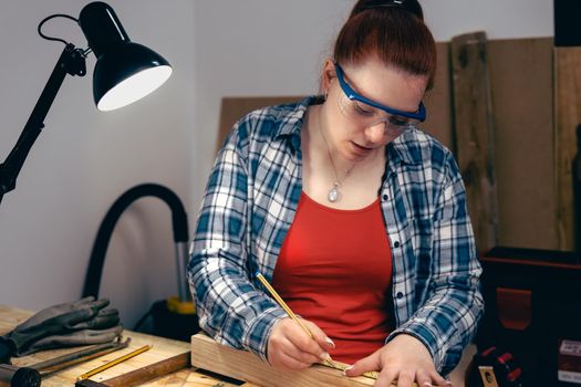 Young carpenter with red hair, working on wood design in a small carpentry workshop, dressed in blue checked shirt and red t-shirt. Young businesswoman working by hand on a piece of wood and designing new furniture for the house. Warm light indoors, background with wooden slats. Horizntal.