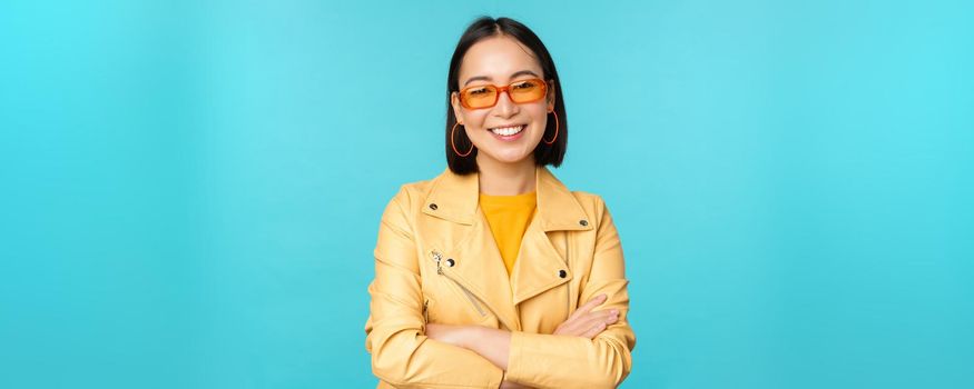 Stylish korean woman in sunglasses and yellow jacket, cross arms, smiling and looking confident at camera, standing against blue background.