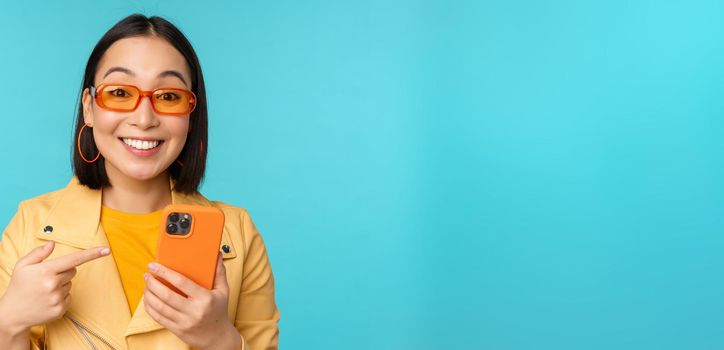 Enthusiastic asian girl in stylish sunglasses, pointing finger at smartphone, showing mobile phone, standing over blue background.