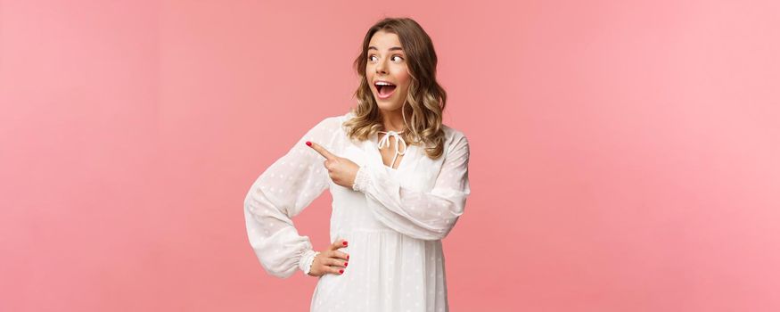 Excited and amazed blond caucasian girl in kawaii dress, open mouth gasping in awe, pointing and looking left with excitement and astonishement, see great beauty product, pink background.