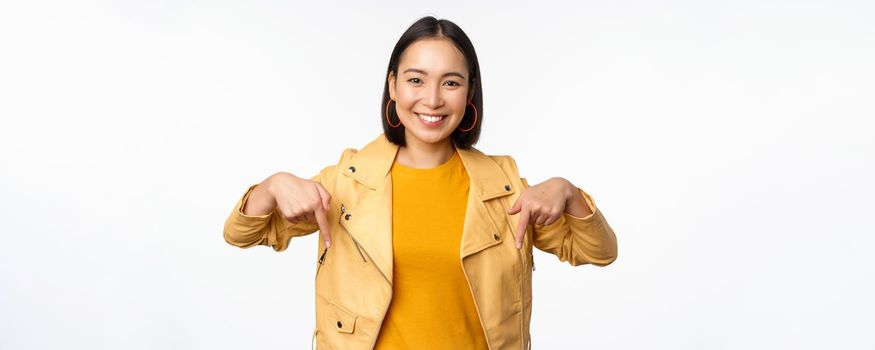 Portrait of happy smiling asian girl, pointing fingers down and showing logo, demonstrating banner, standing in yellow jacket against white background. Copy space